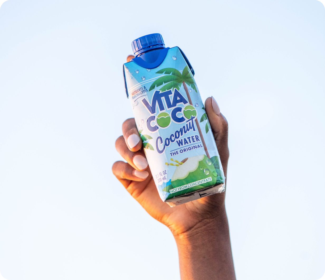 Hand holding a carton of Vita Coco coconut water against a clear sky.